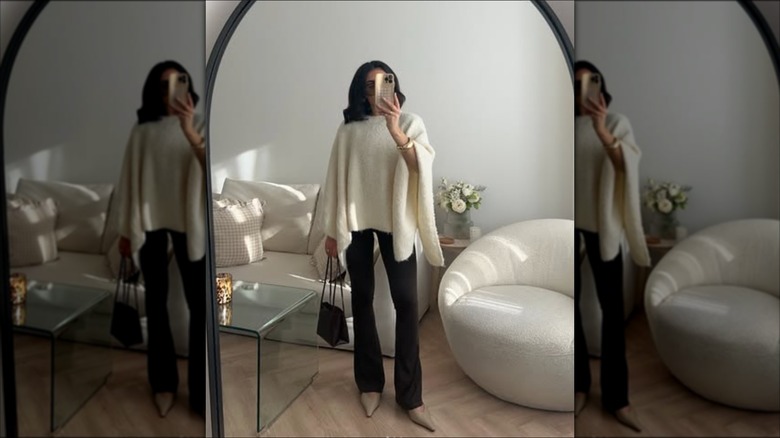 A woman taking a mirror selfie of her outfit with a white knit poncho over dark pants