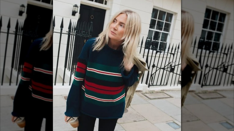 A blonde woman leaning in the street whiel wearing a chunky sweater with red and green stripes