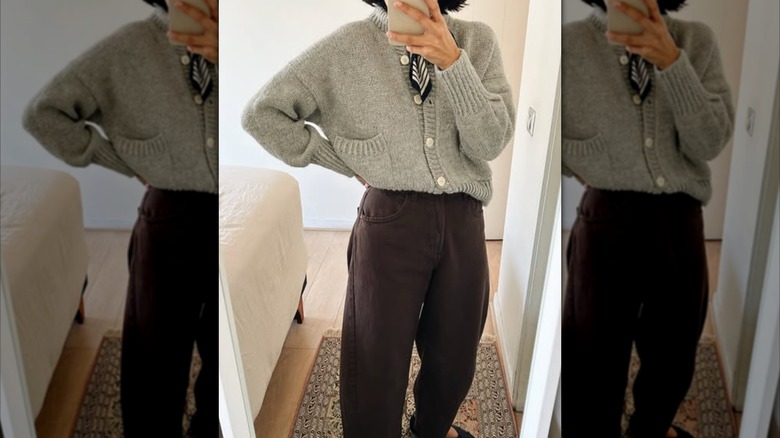 A woman taking a mirror selfie of her outfit with a gray sweater tucked into brown pants