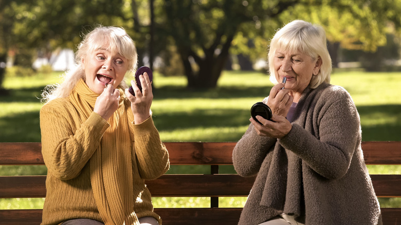 Two older women sat on a bench applying makeup