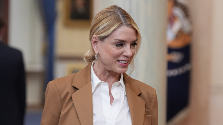 Pam Bondi walking and looking down while smiling, and wearing a tan coat