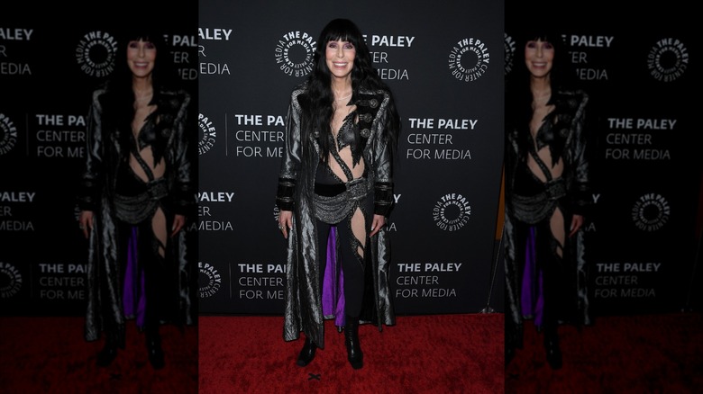 Cher smiles wearing a black bodysuit and a gray sailor-style coat