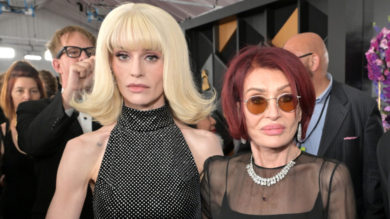 Kelly Osbourne wearing a black dress stands next to Sharon Osbourne wearing sunglasses