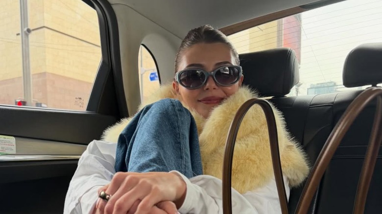 Olivia Jade poses in a car, holding her leg and wearing a fluffy yellow scarf