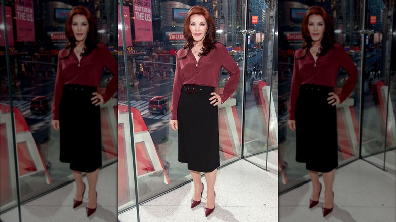 Priscilla Presley wearing a red button-up and black skirt while posing in front of a city window