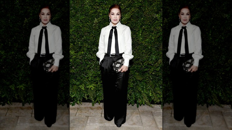 Priscilla Presley posing in front of a hedge wall while wearing a white button-up and satin trousers