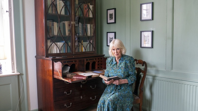 Queen Camilla reading a book while sitting by a bookshelf