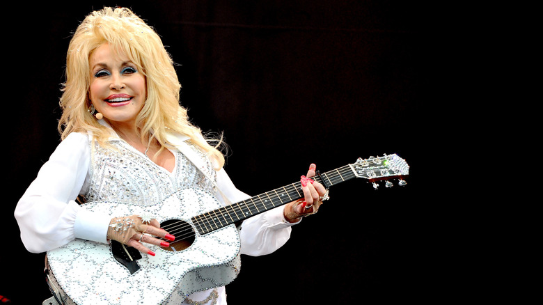 Dolly Parton playing guitar on stage in white with a white guitar