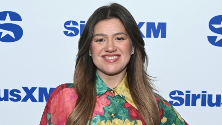 Kelly Clarkson smiling in front of a white background with blue writing