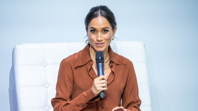 Meghan Markle holding a microphone in a brown blouse