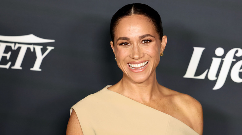 Meghan Markle smiling in a one-shoulder cream dress with her hair slicked back