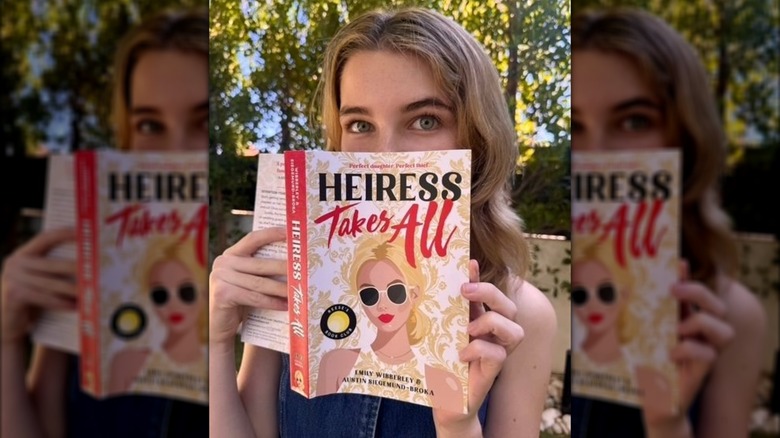 Raegan Revord peeking over a book called "Heiress Takes All"