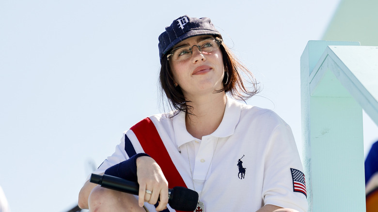 Billie Eilish looking up with a small smile while leaning her arm on her knee in 2024