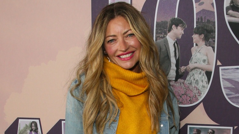 Rebecca Gayheart smiling in front of a pink background while wearing a mustard yellow sweater and denim jacket