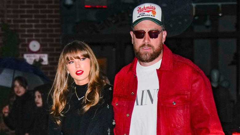 Travis Kelce in sunglasses, a hat and a red suit while Taylor Swift walks beside him in a black dress