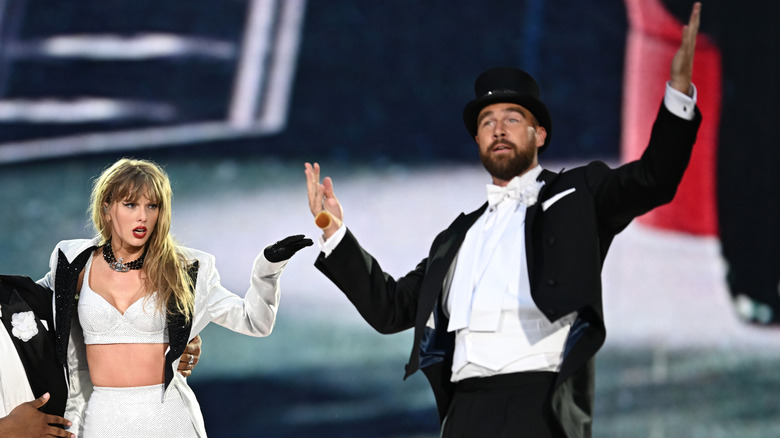 Taylor Swift in a white set and jacket with Travis Kelce in a tuxedo on stage