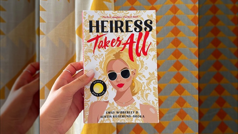A hand holding Heiress Takes All by Emily Wibberley and Austin Siegemund-Broka