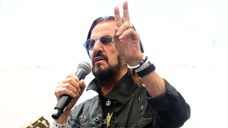 Ringo Starr speaking into a microphone giving a peace sign