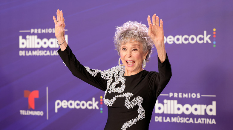 Rita Moreno smiles with her arms in the air wearing an embellished black dress