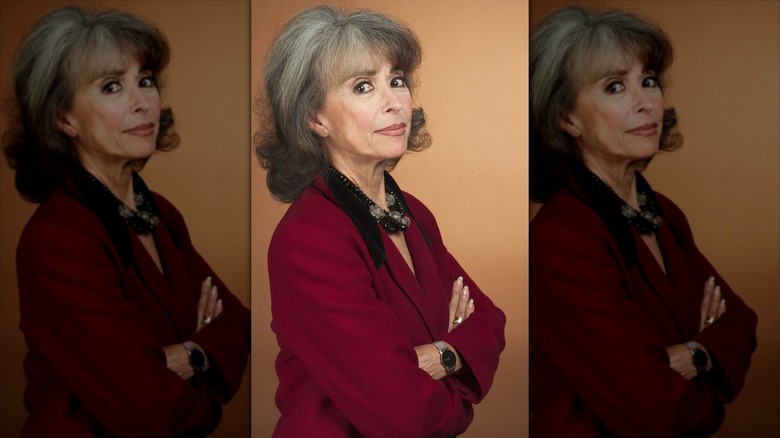 Rita Moreno poses with long gray hair wearing a red blazer