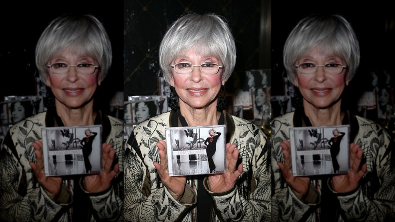 Rita Moreno smiles holding her album wearing white-framed glasses
