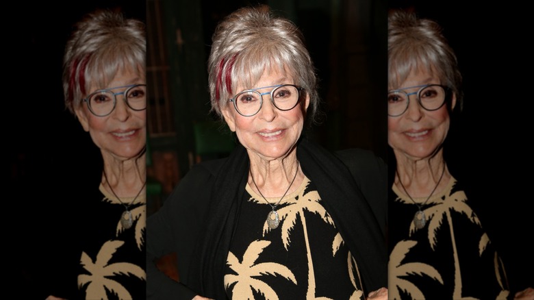 Rita Moreno smiles wearing round glasses and hot pink highlights in her hair