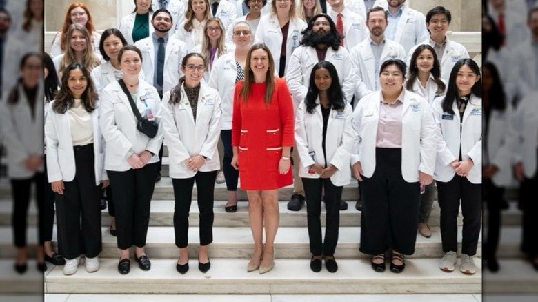 Sarah Huckabee Sanders in a red shift dress and surrounding by medical students in white lab coats on marble steps