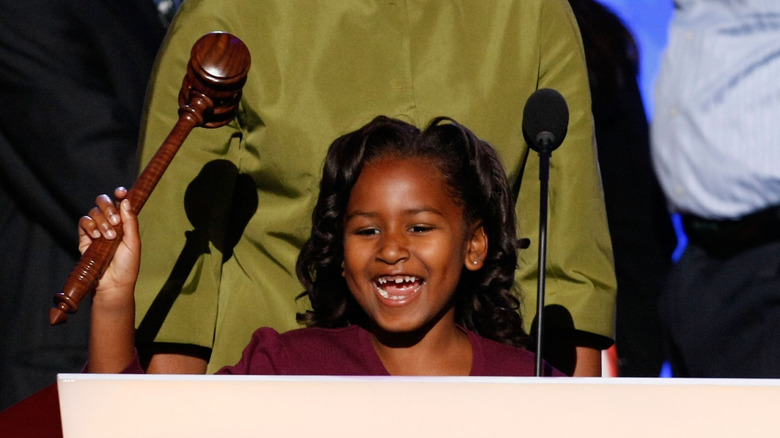 Sasha Obama smiles behind a microphone and holds a gavel