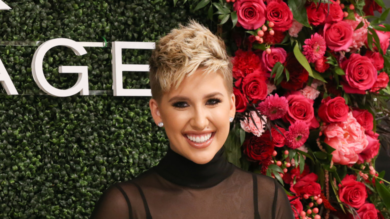 Savannah Chrisley with a pixie haircut, smiling on a red carpet in front of red flowers