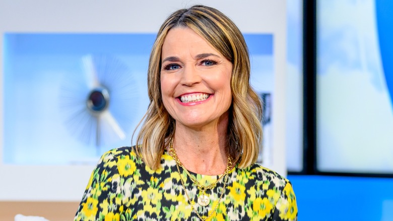 Savannah Guthrie smiling in a TV studio wearing a yellow, white, and black floral print top