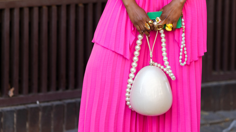 Close-up of a fashion week guest holding an egg-shaped purse with pearl chain against a bright pink dress