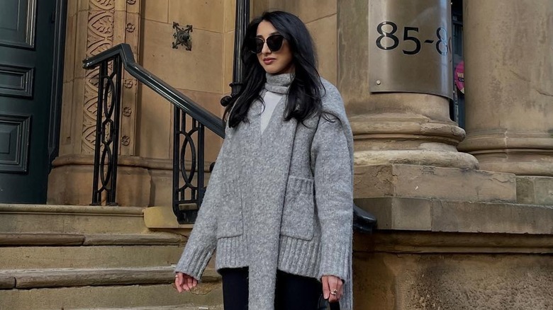 A woman posing while wearing sunglasses and a gray cardigan with an attached scarf