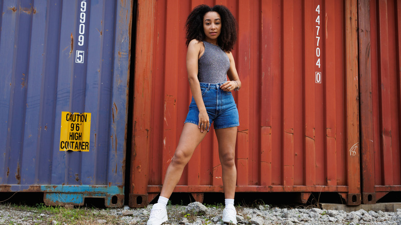 Young woman with curly hair posing in front of cargo containers while wearing high-waisted, tight-fit denim shorts