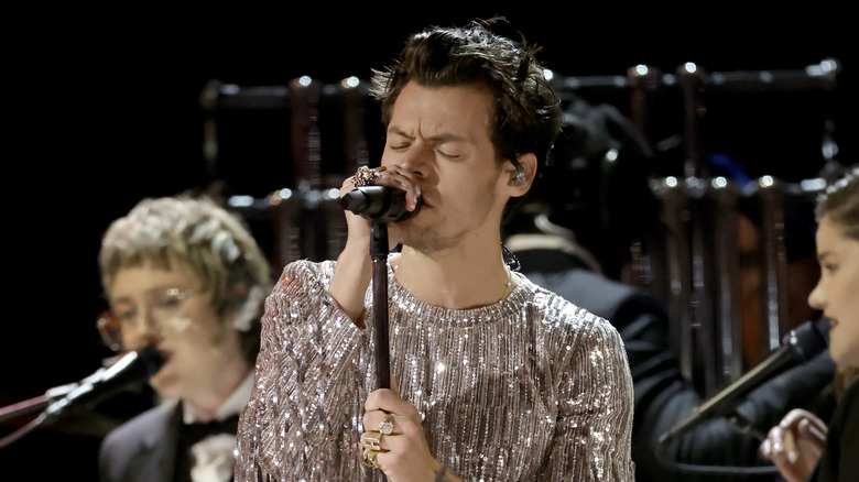 Harry Styles singing into a microphone on stage in a sparkly silver outfit
