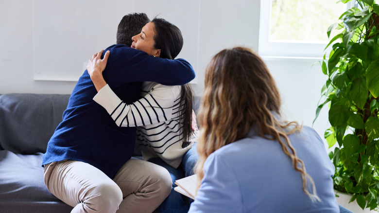 Couple embracing each other while sitting on sofa in front of female psychotherapist at doctor's office after counseling session