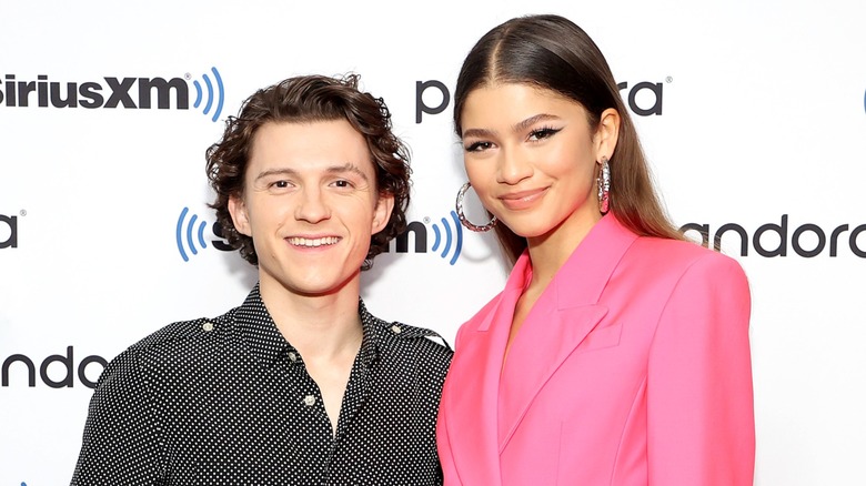 Tom Holland smiling alongside Zendaya on the red carpet in front of a white background