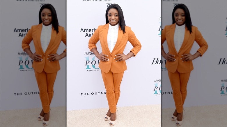Simone Biles posing with her hands on her hips in a burnt orange pantsuit