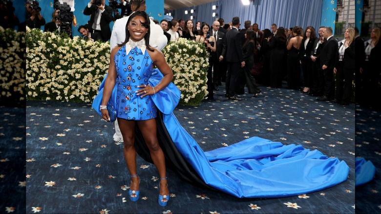 Simone Biles smiling at the 2025 Met Gala while wearing a short blue dress with a long train