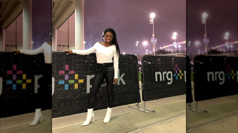Simone Biles posing next to a fence while wearing a white top and leather pants