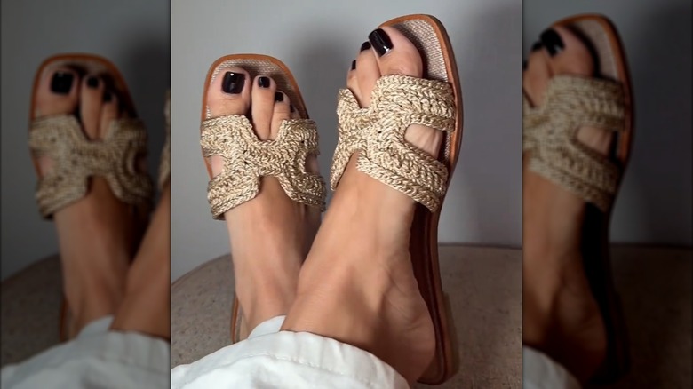 A woman wearing raffia slides with dark nail polish on her toes