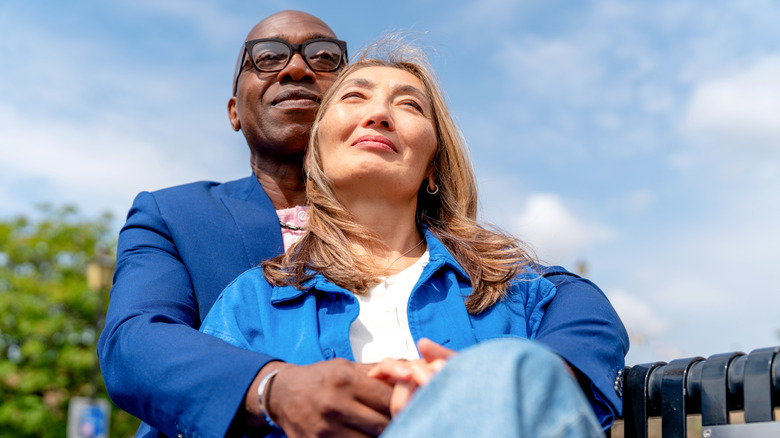 A man and a woman relax on a park bench. They are close to each other, enjoying the sunshine and clear sky
