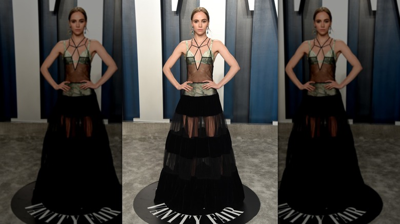 Suki Waterhouse standing with her hands on her hips while wearing a sheer dress over a green two-piece