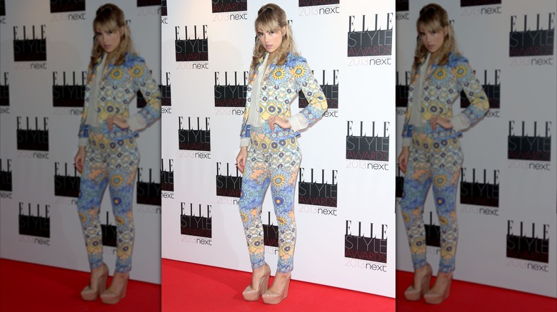 Suki Waterhouse posing with a hand on her hip while wearing a patterned blazer and skinny pants