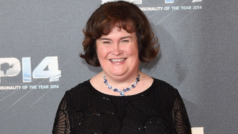 Susan Boyle smiling on the red carpet in 2014 in front of a gray background