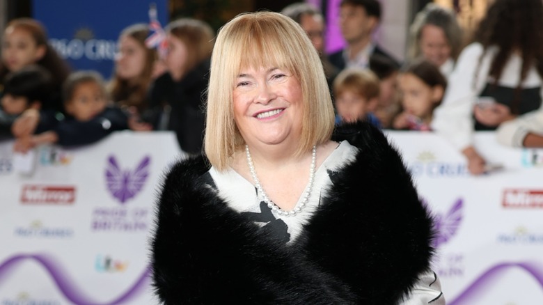 Susan Boyle smiling with a blond bob and bangs on the red carpet in 2025