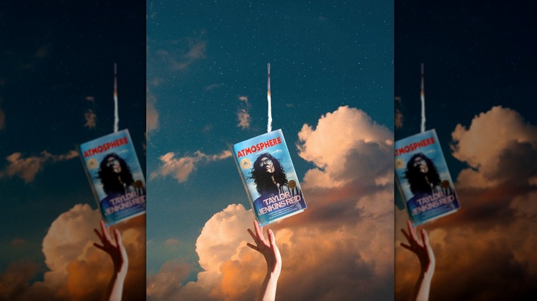 A hand holding a copy of the book "Atmosphere" in front of a rocket launching into the sky