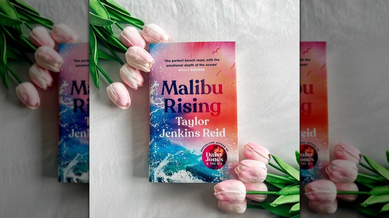 A copy of "Malibu Rising" laying on a white sheet with some pink tulips