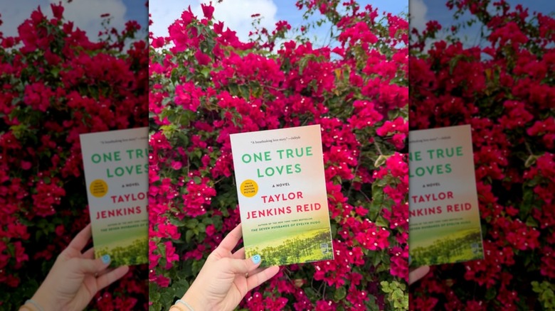 A hand holding up a copy of the book "One True Loves" in front of bush of pink flowers