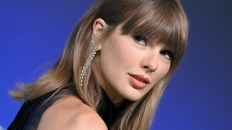 Angled snapshot of Taylor Swift looking over her shoulder while wearing a peach-tinted lip color and dangly earrings in 2026