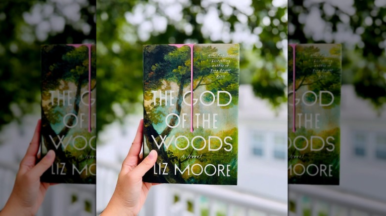 A hand holding up a copy of "The God of the Woods" with a blurry, leafy background behind